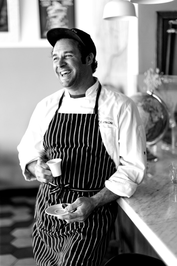 Chef Accursio Lota smiling in an apron while holding a cup of espresso, captured in black and white inside Cori Trattoria.