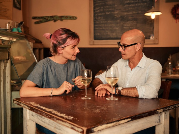 Stanley Tucci chats with a woman over glasses of white wine in a cozy trattoria.