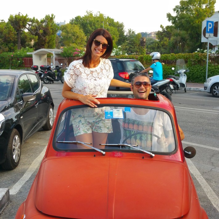 Vincenzo and his wife Suzanne smiling in a vintage red car.