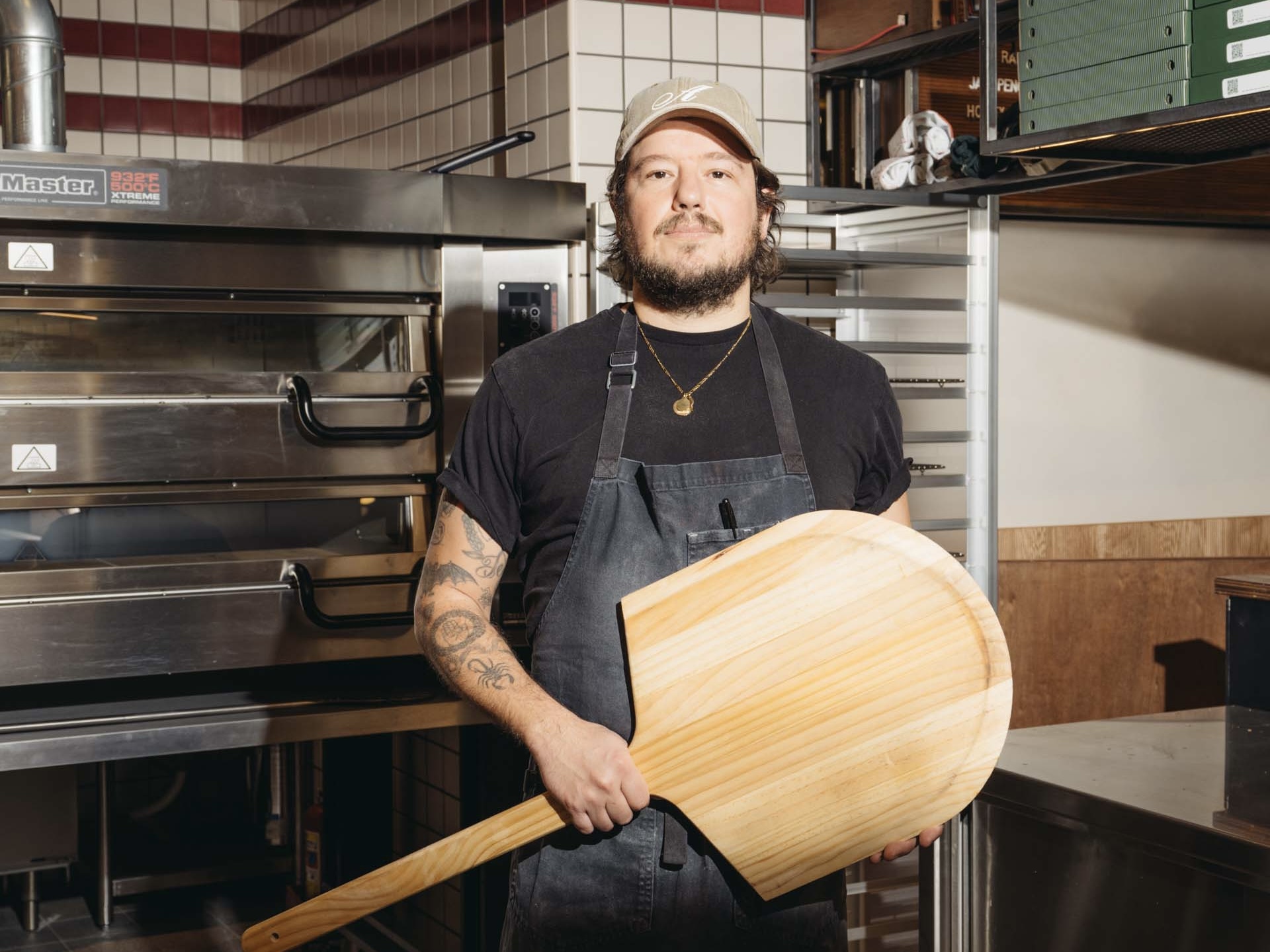 Anthony Falco, author of Pizza Czar, standing in a kitchen wearing a black apron and holding a pizza peel.