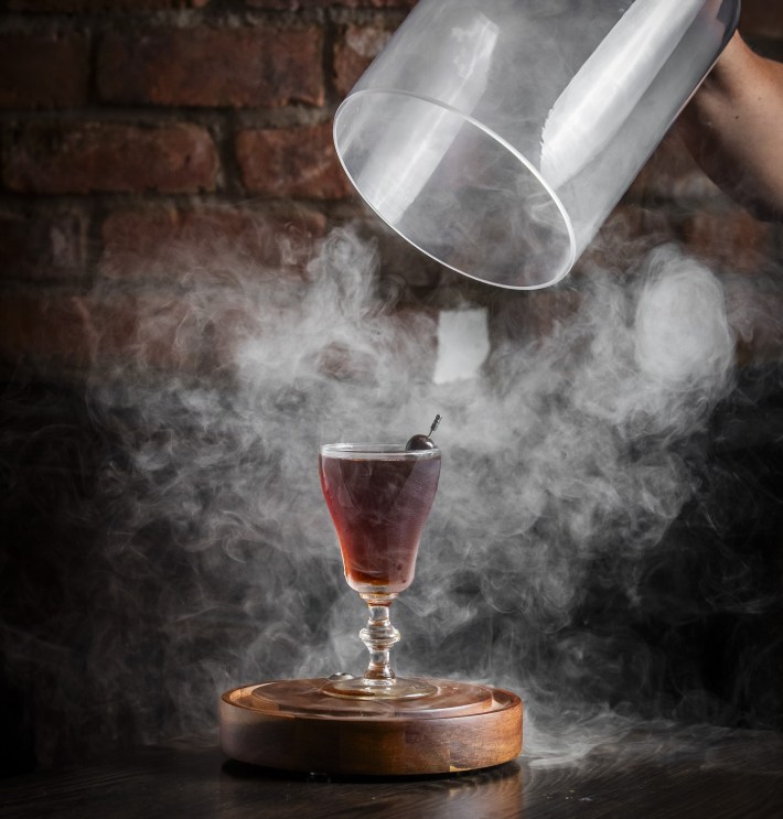 Smoked cocktail being unveiled from a glass lid with swirling vapor