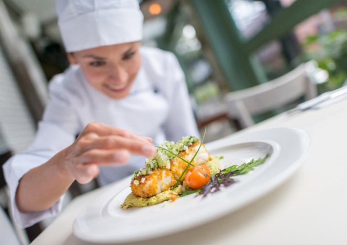 A smiling chef carefully plating a dish topped with herbs and garnishes.