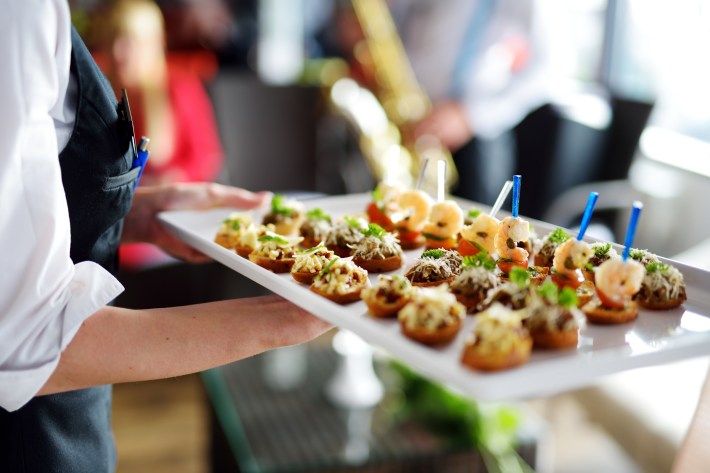 A server holding a tray of assorted seafood and crostini appetizers at an event.