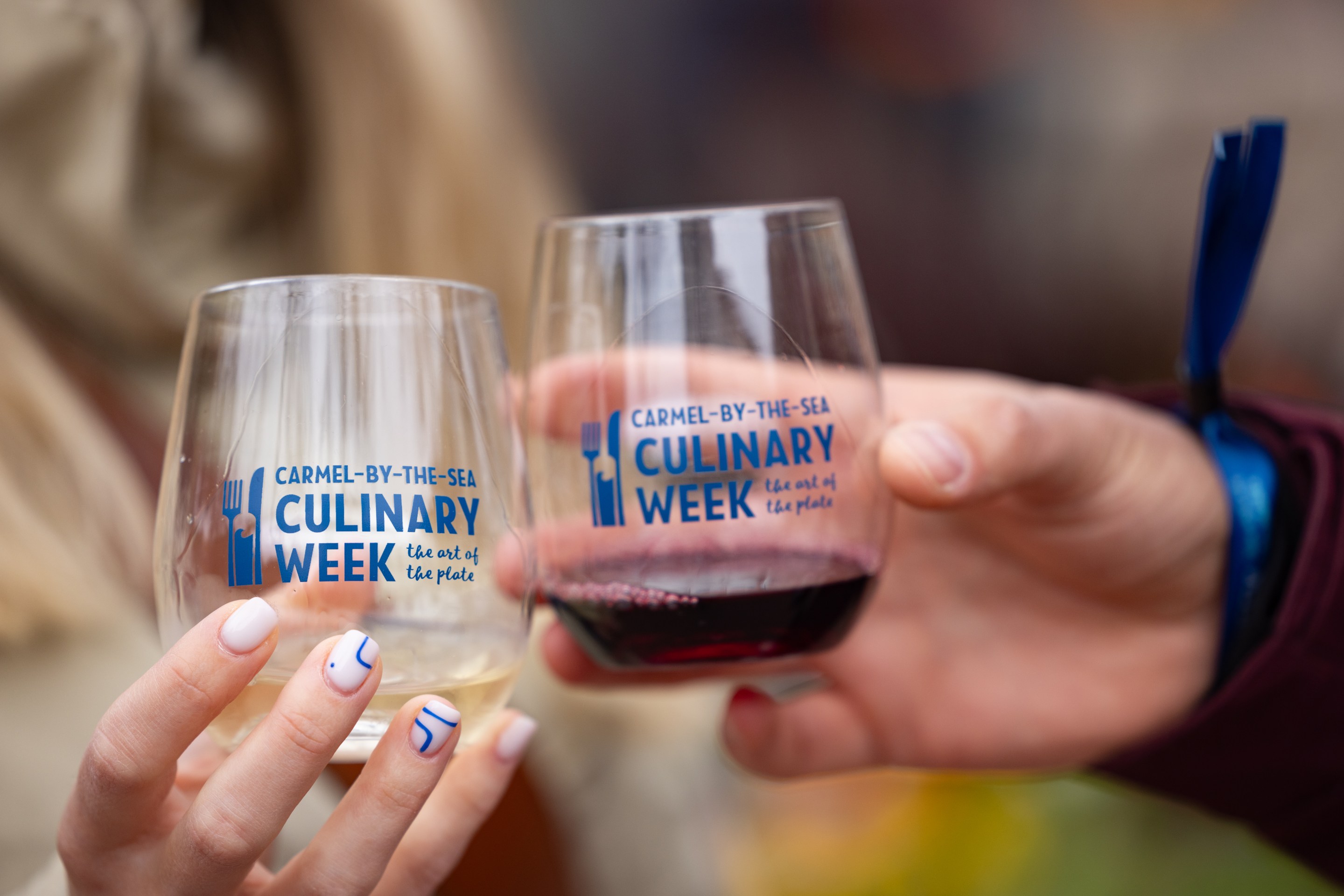 Two people clink wine glasses branded with the Carmel Culinary Week logo.