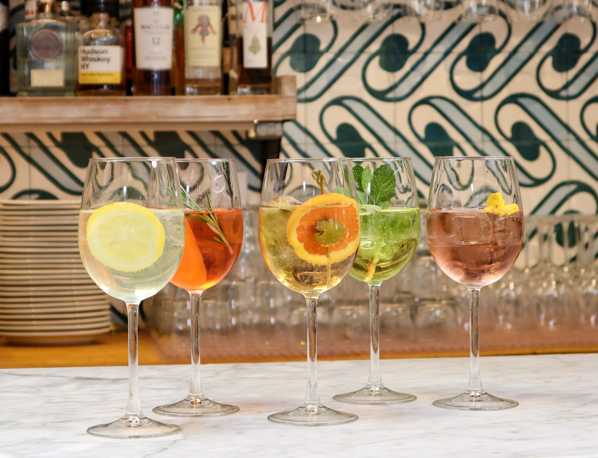 Five colorful spritz cocktails garnished with citrus, herbs, and flowers, displayed at the bar.