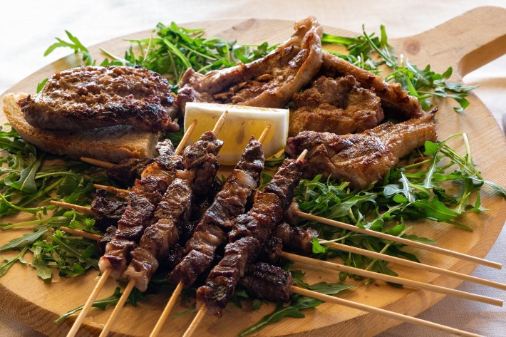 A wooden board with grilled lamb chops, lamb skewers, and greens, garnished with a lemon wedge.