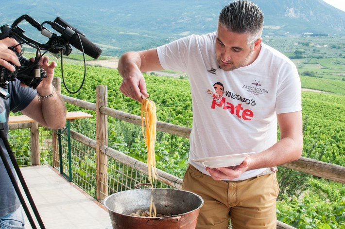 Vincenzo cooking pasta outdoors with a vineyard in the background.