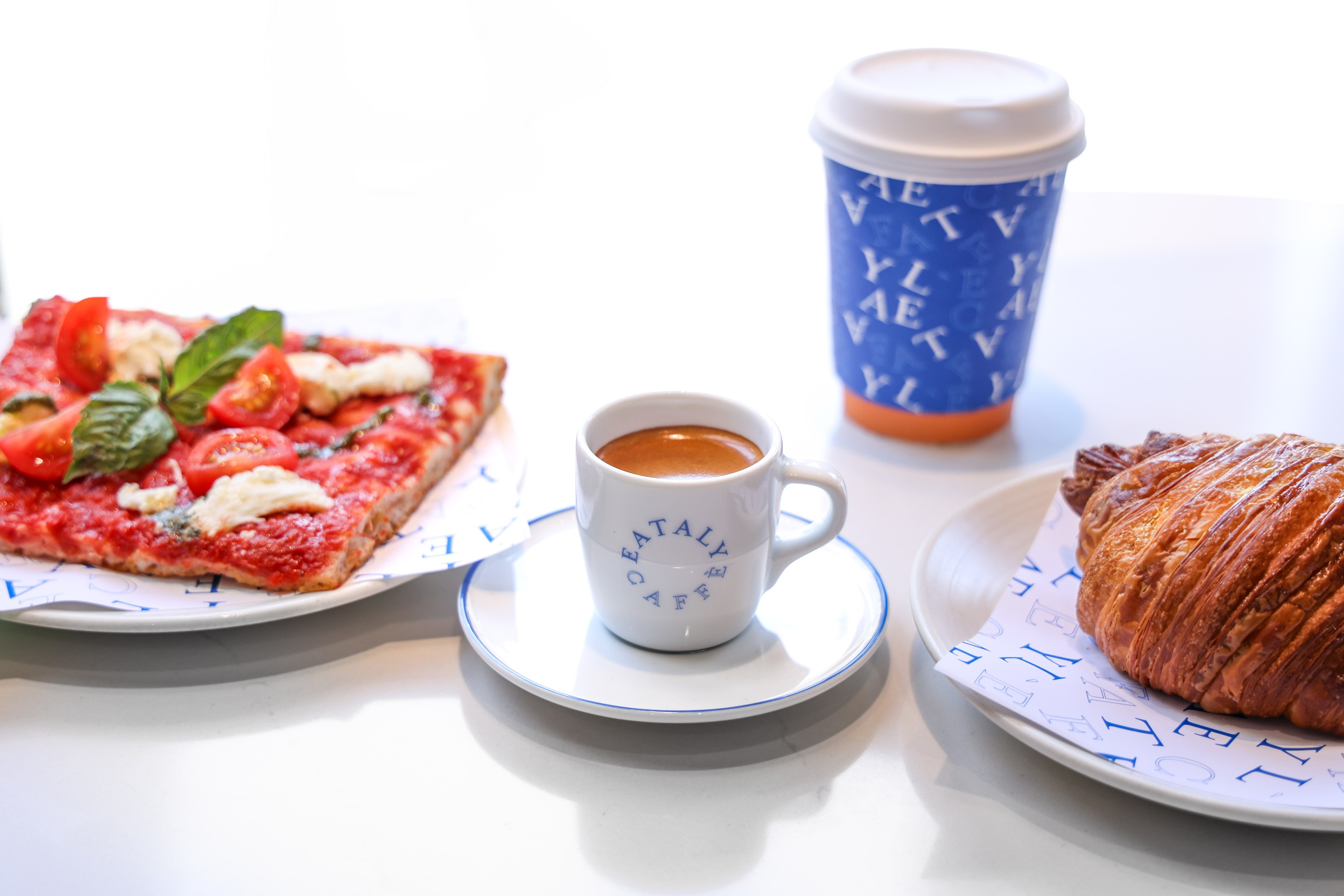 A breakfast spread from Eataly Caffè featuring a slice of focaccia-style pizza with cherry tomatoes and fresh basil, a flaky croissant, a cup of espresso, and a branded blue to-go coffee cup.