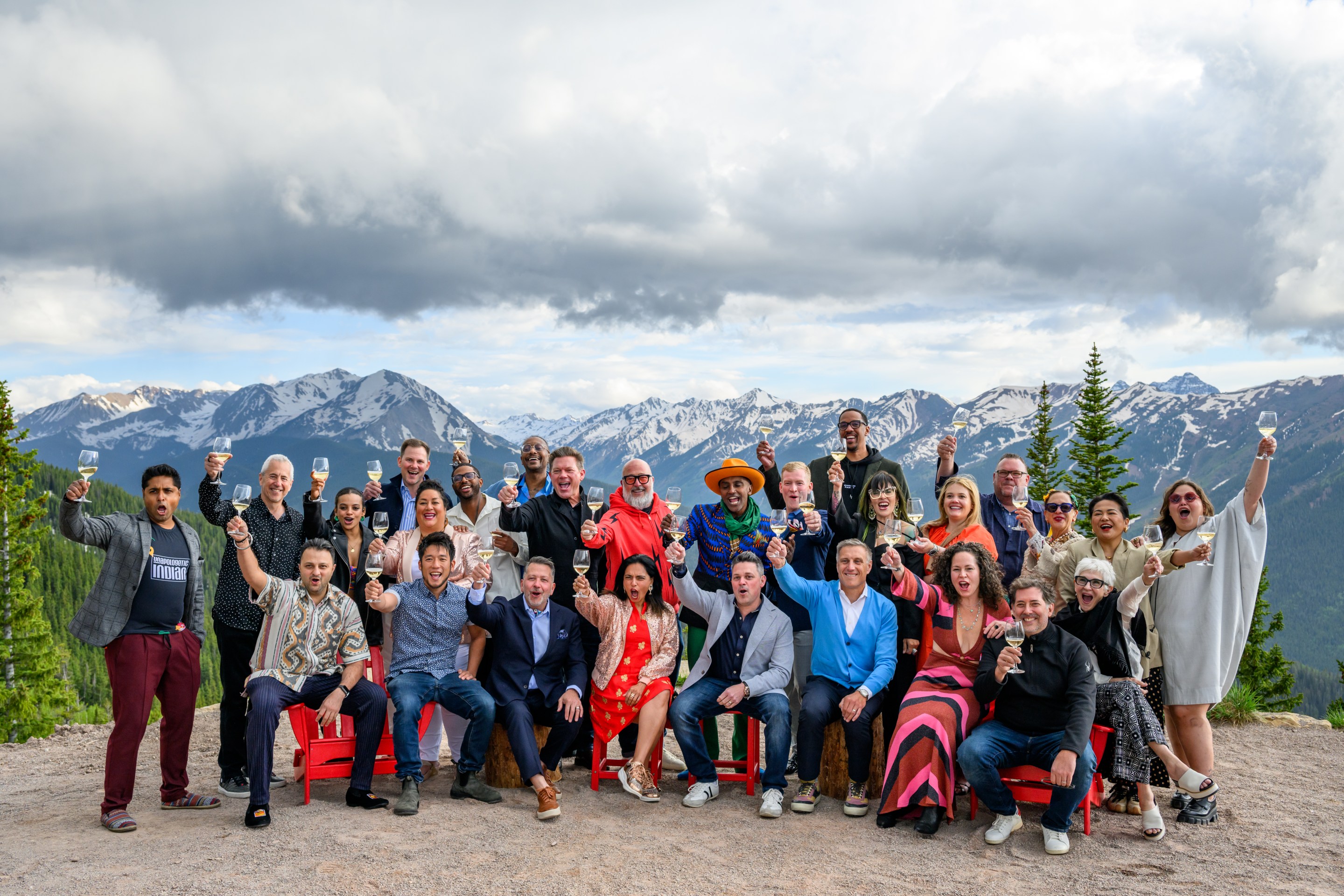 A group of chefs, winemakers, and food industry professionals raise glasses for a toast against a backdrop of snowcapped Rocky Mountains.
