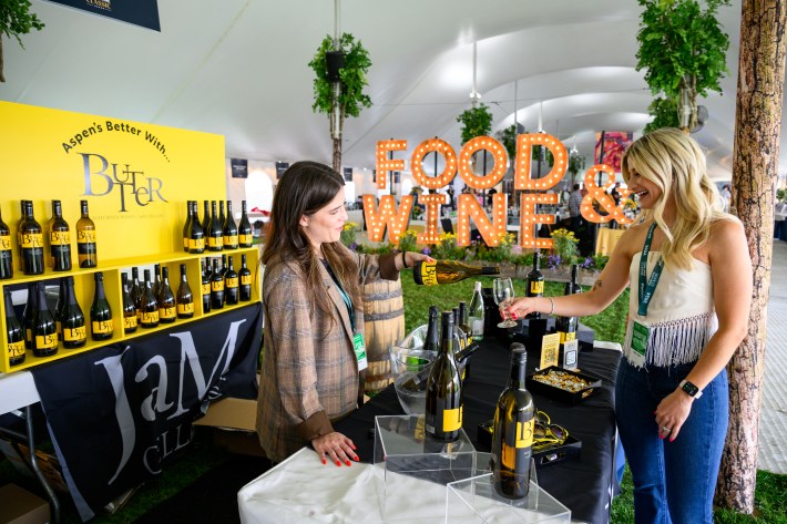 A woman pours wine from a JaM Cellars bottle for a guest at the Food & Wine Classic, with a bright yellow booth and “FOOD & WINE” sign in the background.