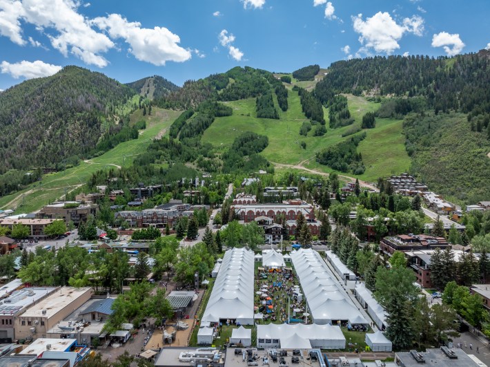 Aerial view of the Food & Wine Classic in Aspen, showcasing rows of white tents set against green ski slopes and the town’s surrounding buildings.