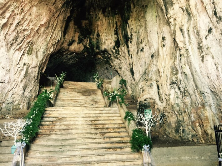 Stone staircase decorated with greenery and white flowers leading into a cave at Santuario Madonna della Grotta in Praia a Mare, Calabria.