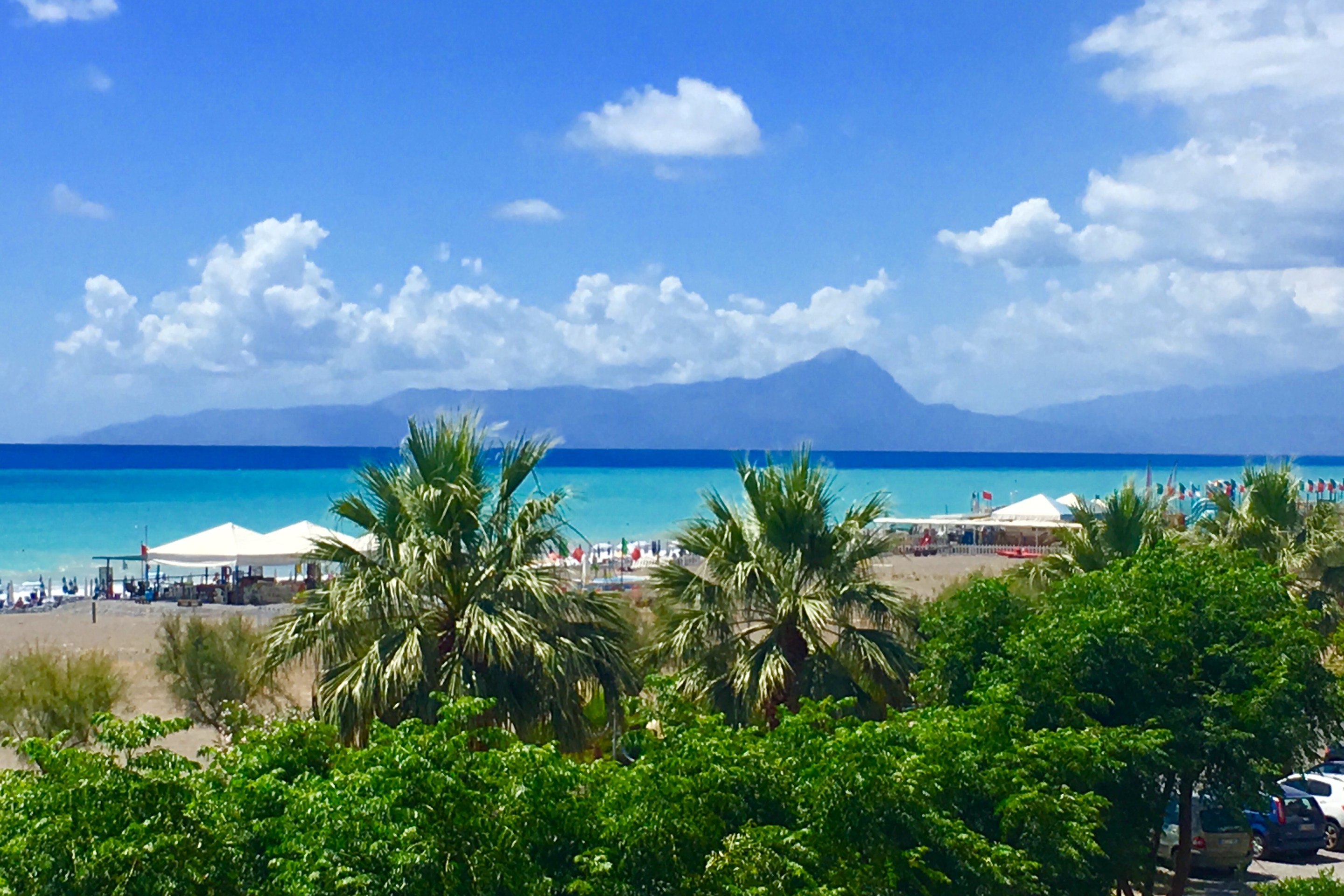 View of Praia a Mare beach with palm trees, turquoise sea, and distant mountains on a sunny summer day in Calabria, Italy.