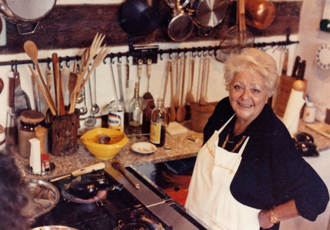 Marcella Hazan smiling at the camera in her Venice kitchen, surrounded by cooking tools and ingredients.