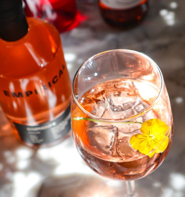 A close-up of a pink spritz cocktail with a yellow flower garnish next to a bottle of Empirical.