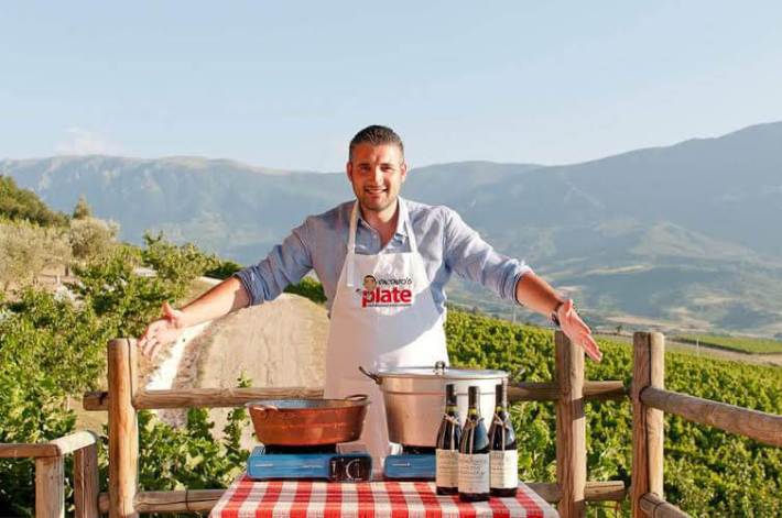 Vincenzo standing behind a table set with cooking pots and wine bottles in the vineyard.