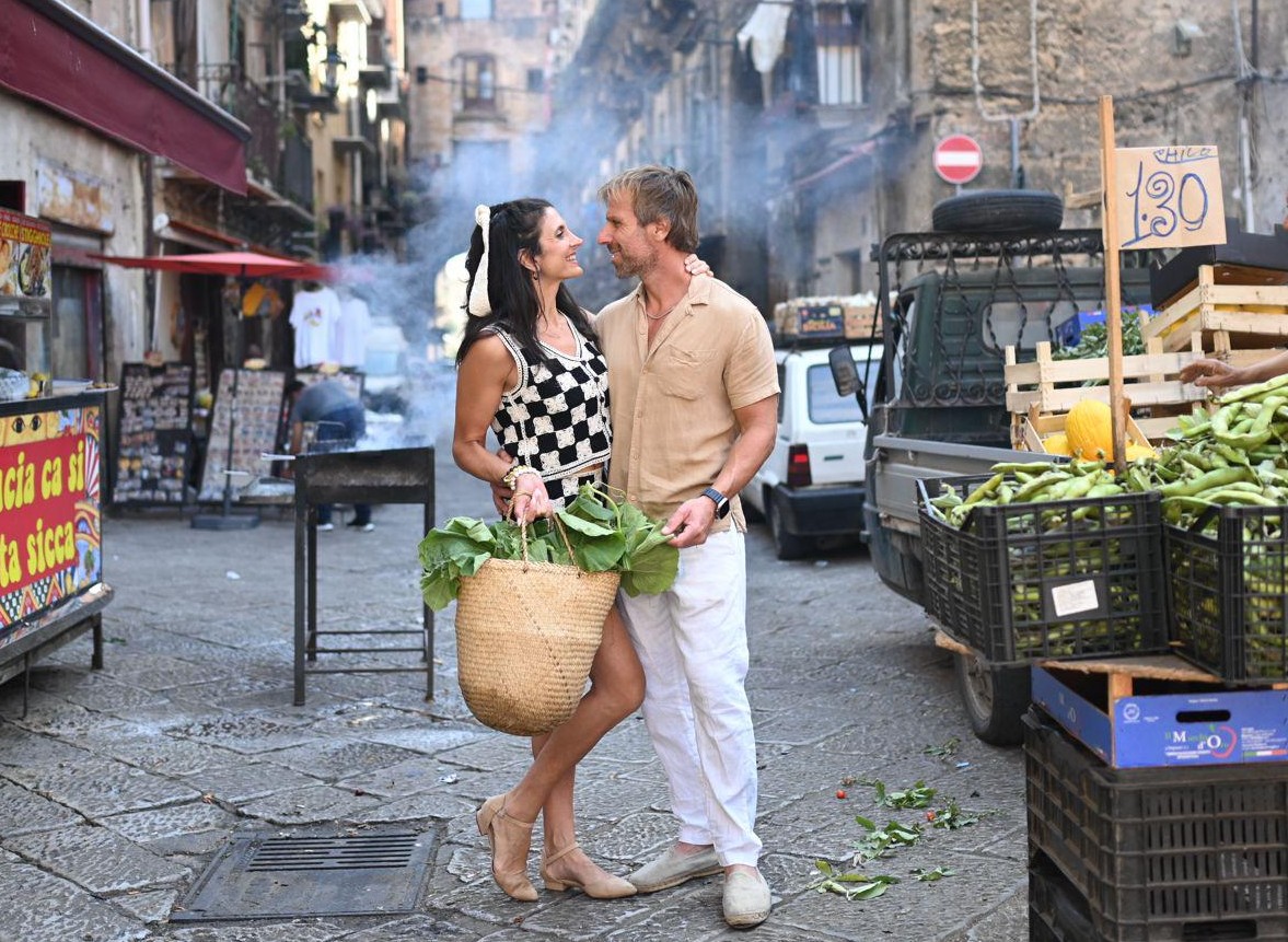 ara and Santi stand close and smiling in a Palermo street market surrounded by produce.