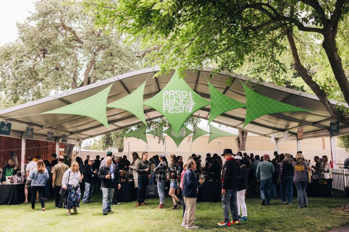 Crowds gather under a large tent decorated with green banners at Paso Wine Fest, sampling wines and enjoying the lively outdoor event.