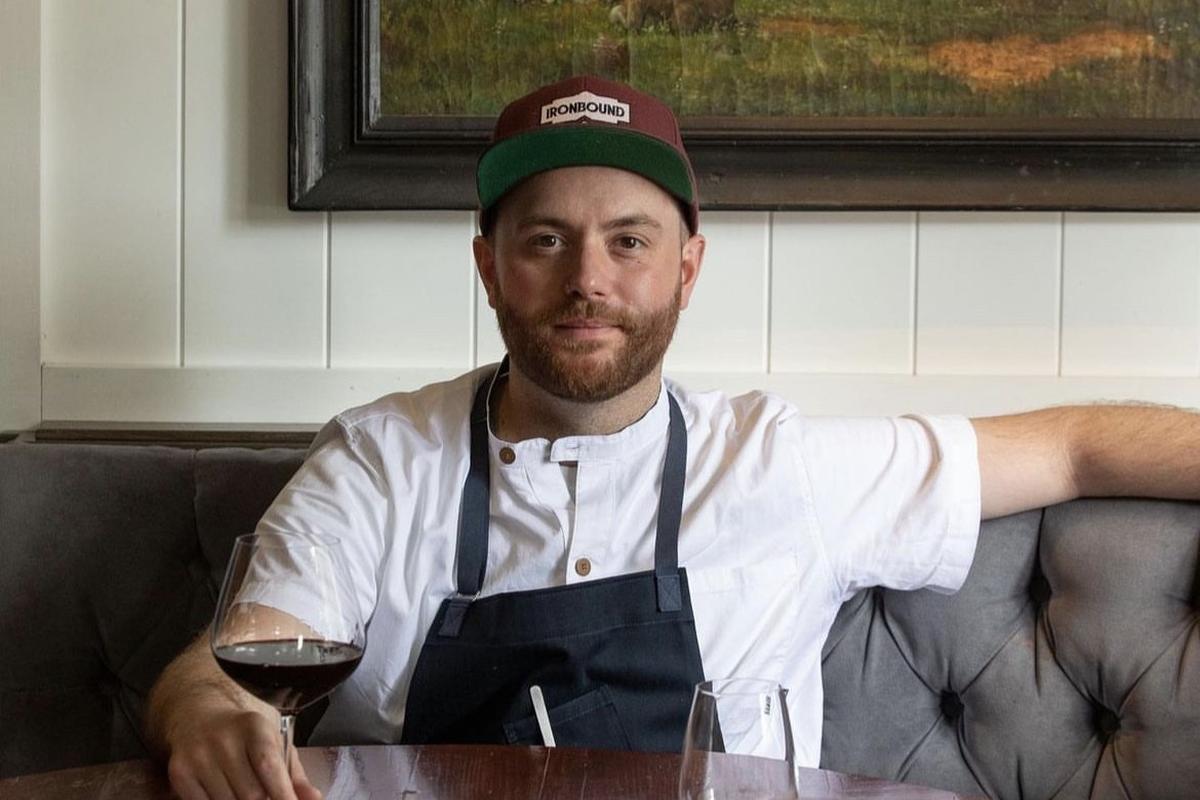 Chef AJ Capella seated in apron and hat, relaxing with a glass of wine.