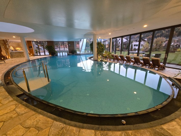 View of indoor pool, lounger chairs lined up for guests, and white resort buildings through the large windows.