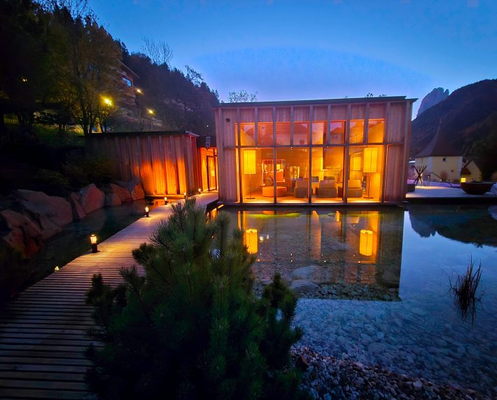 View of lighted Aufguss spa room, with lighted path, still water feature, and hills in the background.