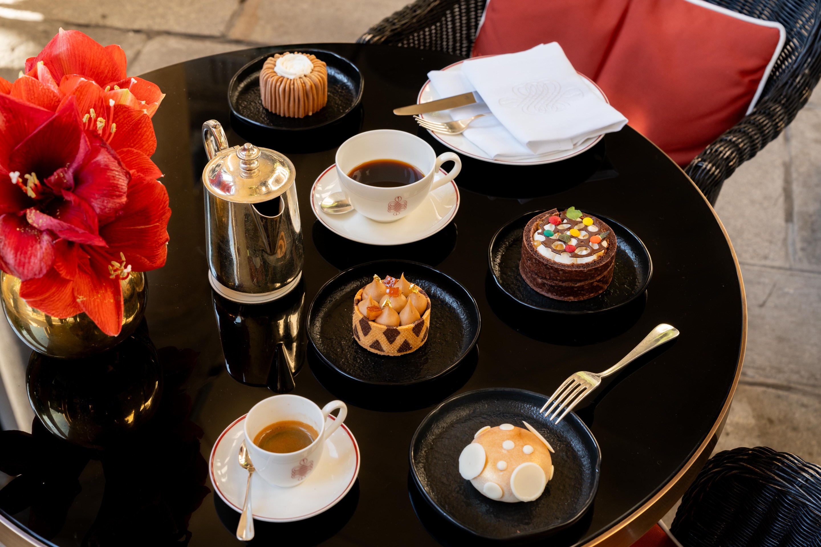 A black table at 10_11 Bar set with espresso, silver coffee pot, and five designer cakes inspired by Ferragamo shoes, with red flowers in a gold vase.