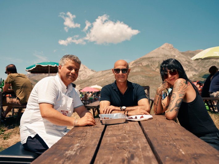 Tucci with restaurant owner and food journalist at table with mountains and blue sky in background.