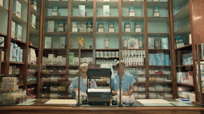Two candy store workers with white hair nets and blue uniforms, and Confetti boxes all around them in shelves.