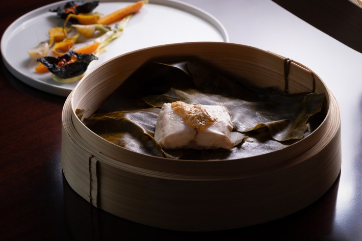 Steamed branzino inside a bamboo steamer, with a plate of vegetables in the background