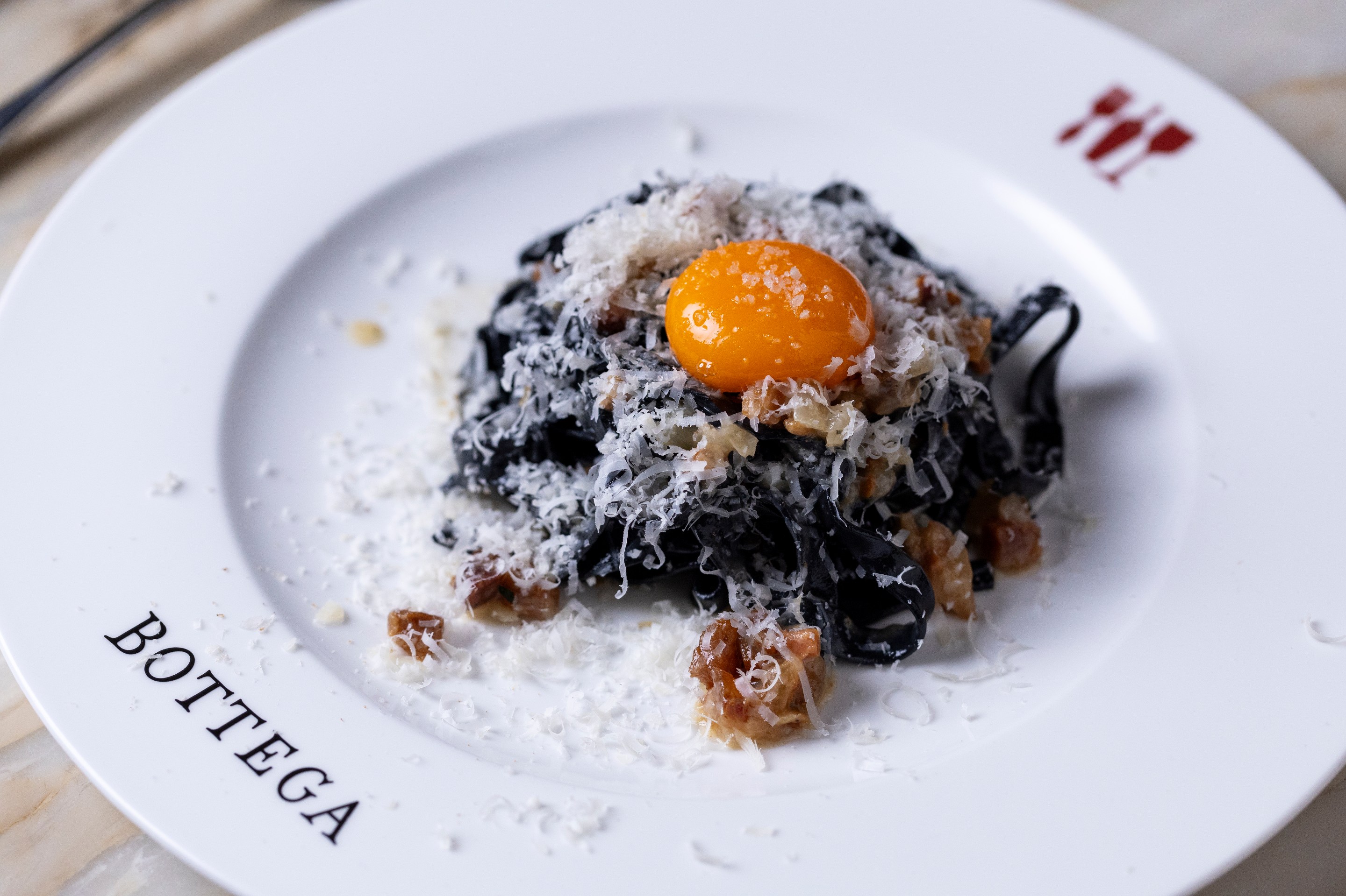 Black pasta topped with an egg yolk and shaved cheese on a white Bottega plate