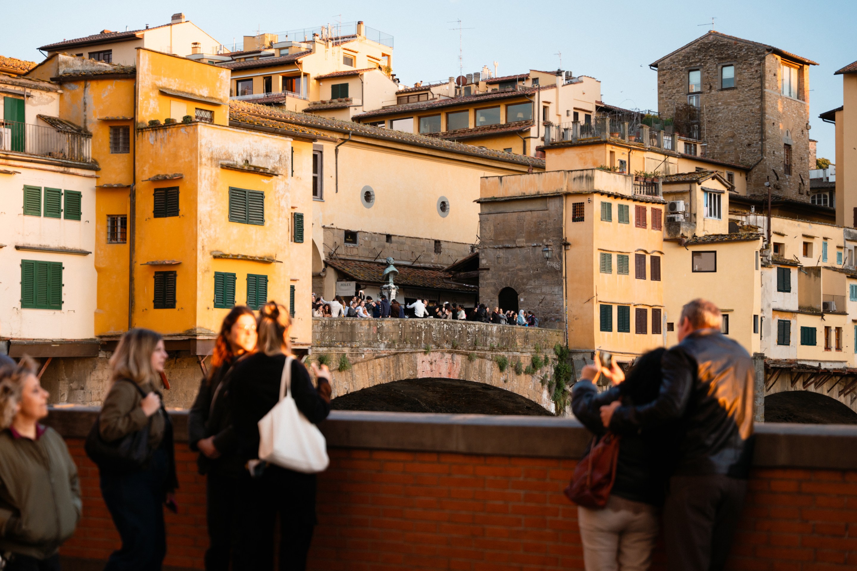 Tourists gathered on a walkway overlooking the Ponte Vecchio and yellow historic buildings in Florence
