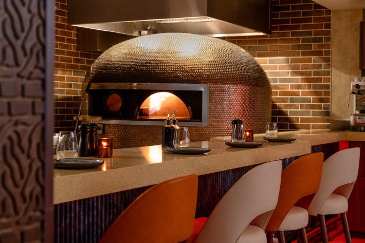 Copper-toned pizza oven glowing with fire behind a counter setup.