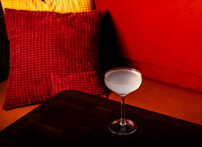 White cocktail in a glass on a dark wood table with red cushions in the background