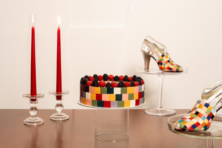 Multi-colored checkerboard round cake topped with berries, next to same-patterned heels and two tall, red candles.