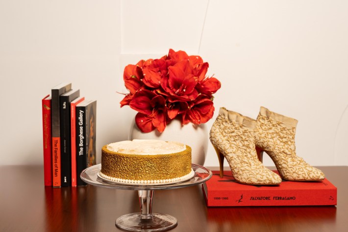 Gold patterned bootie-styled heels and matching round cake, next to red flowers and stacks of books.