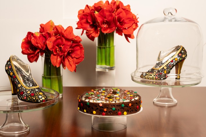 Multi-colored dotted round cake with matching heels, both on cake platters, next to two bouquets of red flowers.