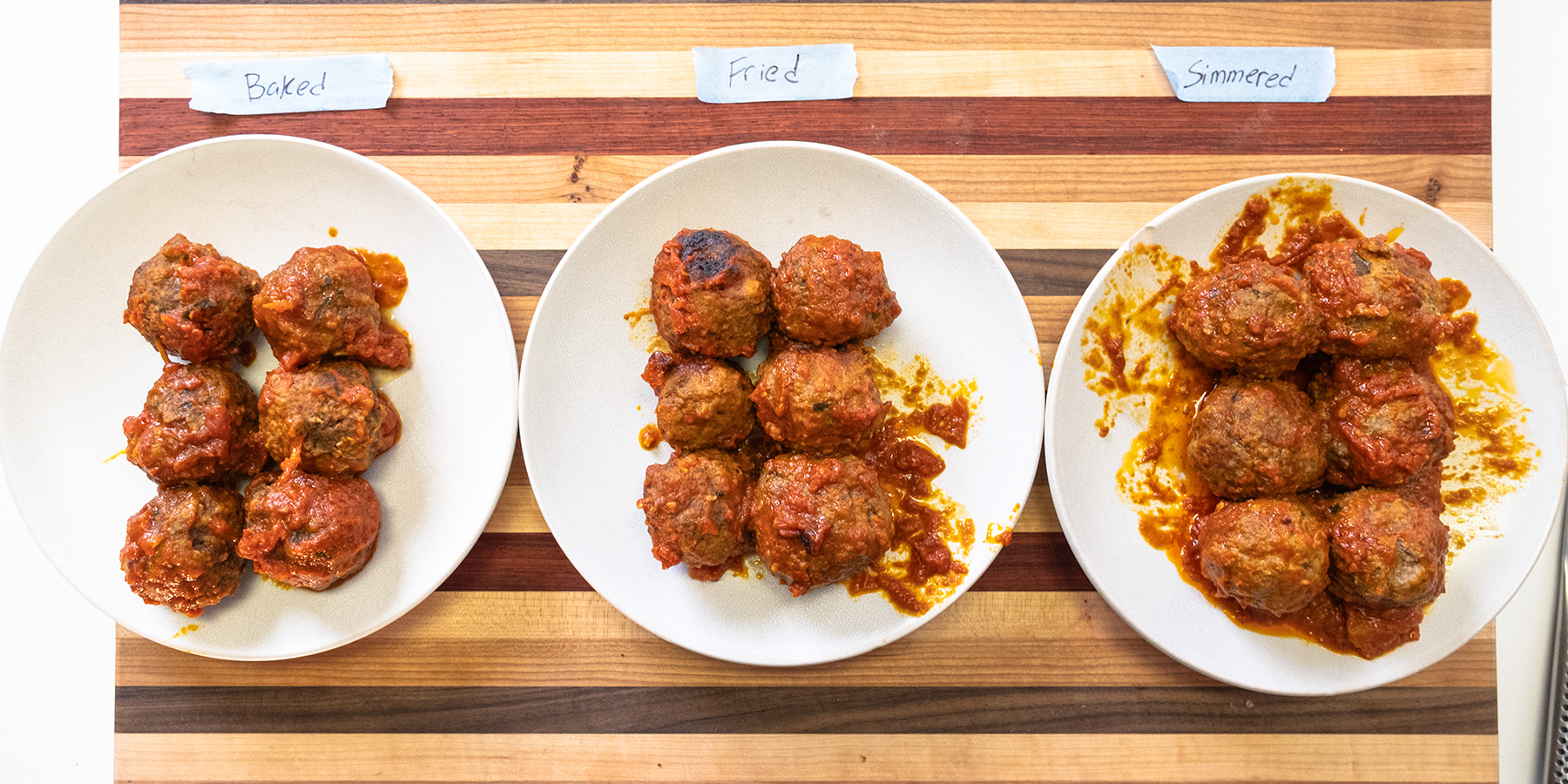 Three plates of meatballs prepared using baked, fried, and simmered methods, labeled on a wooden cutting board.