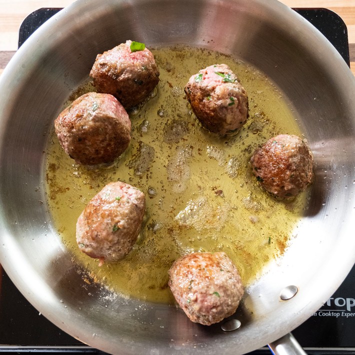 Meatballs frying in olive oil in a stainless steel pan.