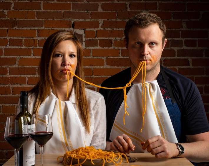 Jackson and Melissa Kalb sharing a plate of spaghetti with red wine in front of a brick wall