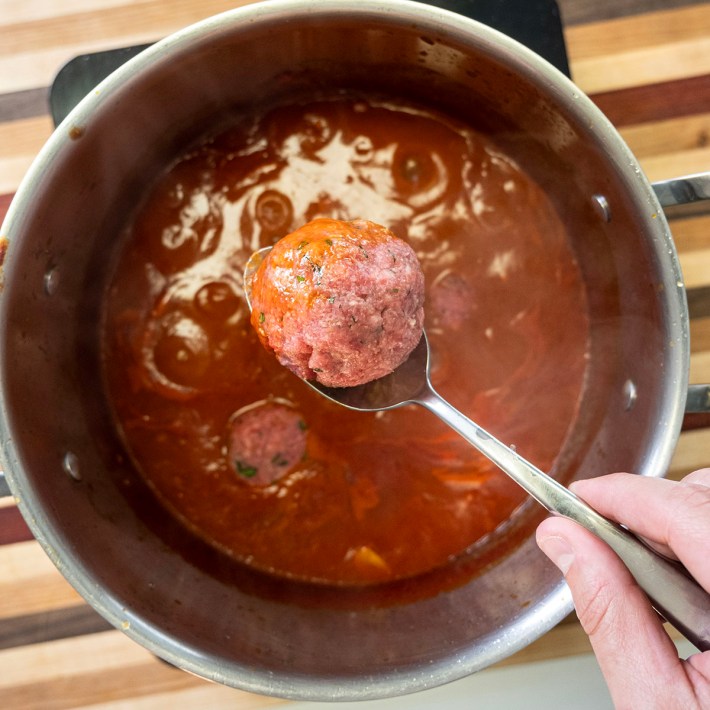 Raw meatball being lowered into a pot of tomato sauce to simmer