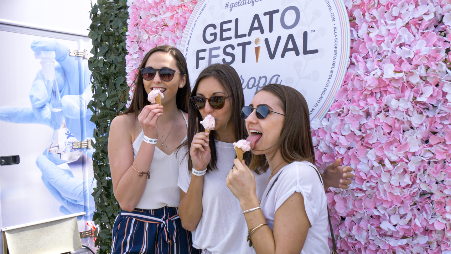 Three women enjoying gelato cones in front of a Gelato Festival sign with a floral backdrop.