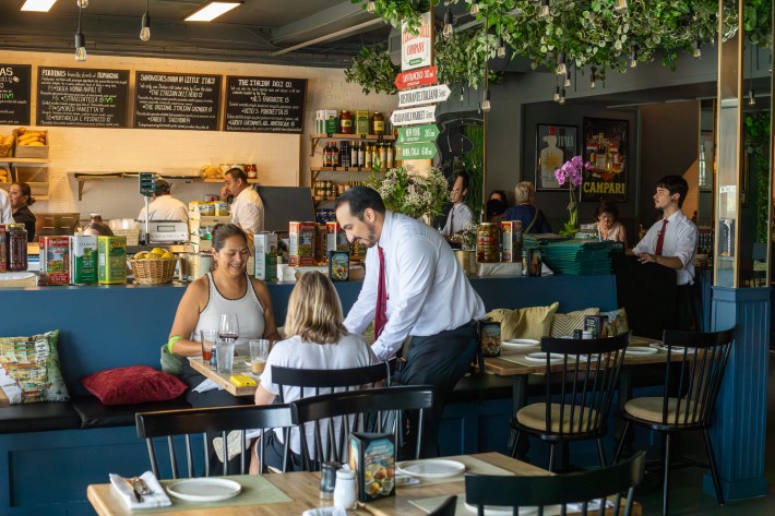 Guests dining inside The Italian Deli Co. in Pasadena with staff serving and chefs preparing food behind the counter.
