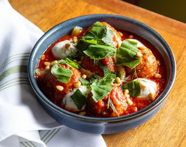 Meatballs topped with pine nuts and basil in a red sauce, in a blue bowl atop a wooden table.