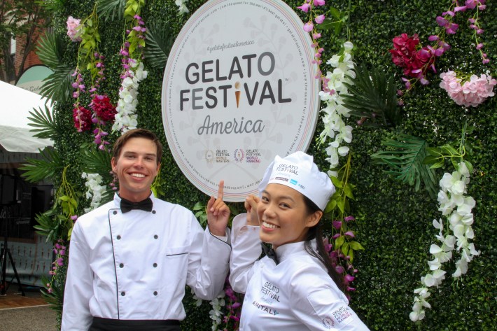 Two smiling chefs in uniform pointing at the Gelato Festival America sign surrounded by flowers.