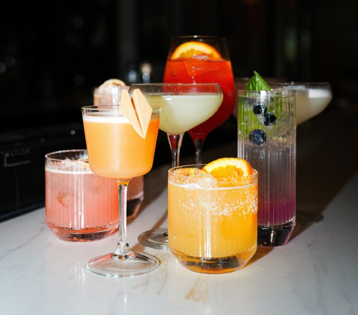 A collection of colorful cocktails garnished with citrus, herbs, and berries displayed on a marble bar.
