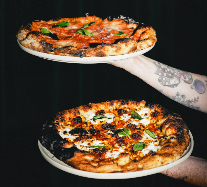 Two Bacarosa pizzas are being held by two arms with tattoos on a black background.