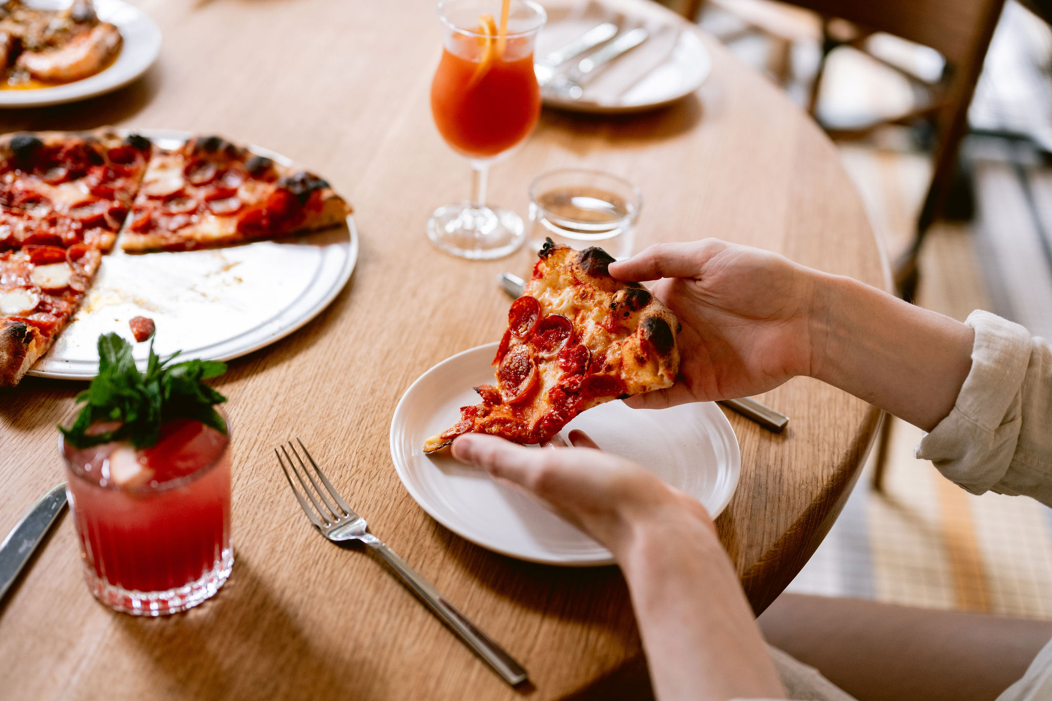 A slice of Flour + Water pizza on a plate lifted by hands with a garnished drink