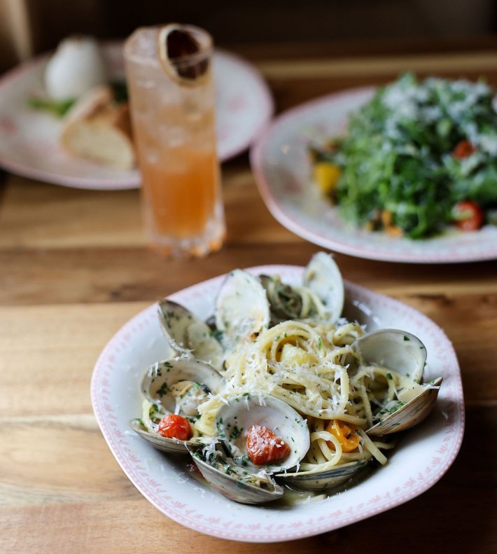 Clam linguine at Allora, set in pasta with tomatos, next to a drink on a wooden table.