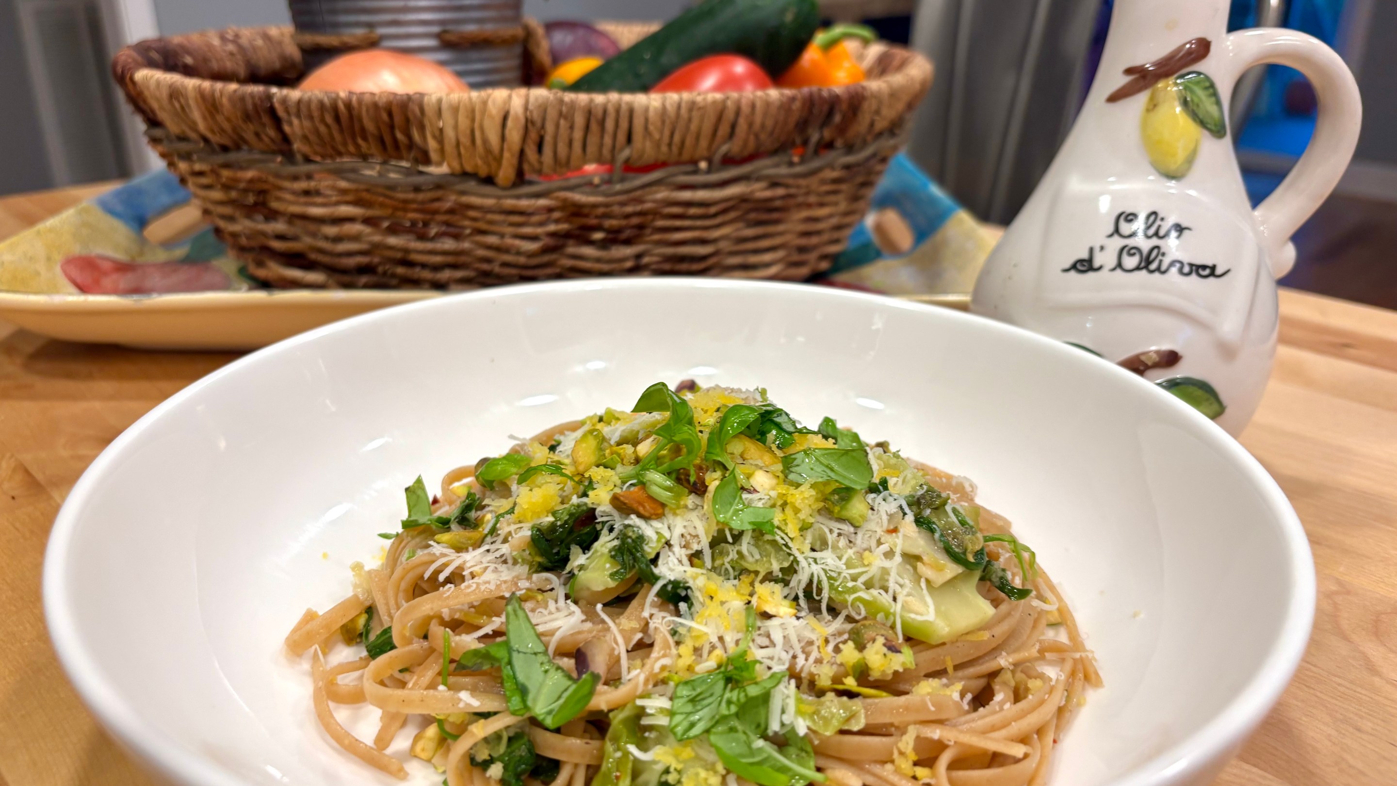 A bowl of whole grain pasta with vegetables, pistachios, and cheese on a kitchen table with a basket of produce in the background.