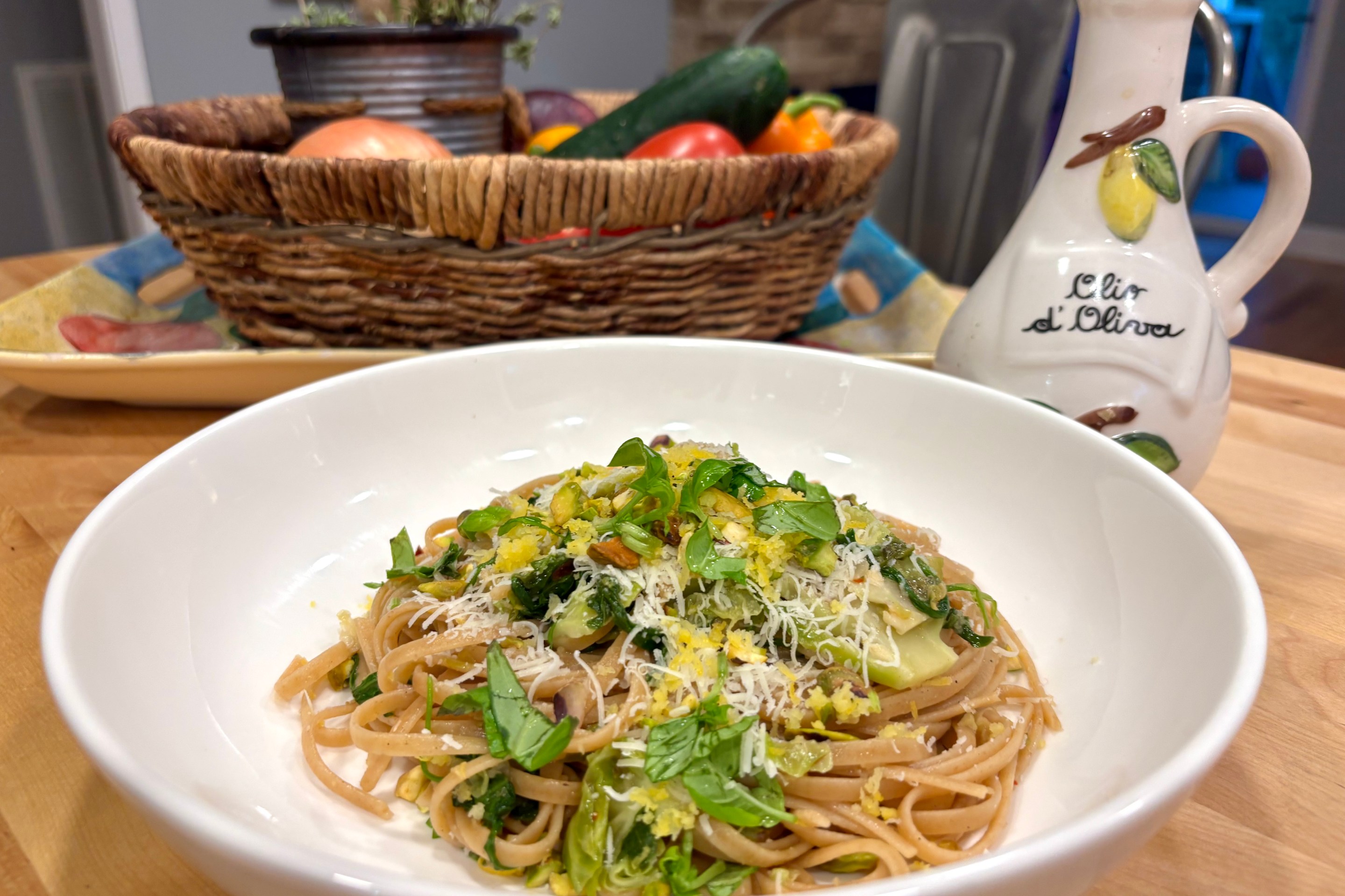A bowl of whole grain pasta with vegetables, pistachios, and cheese on a kitchen table with a basket of produce in the background.